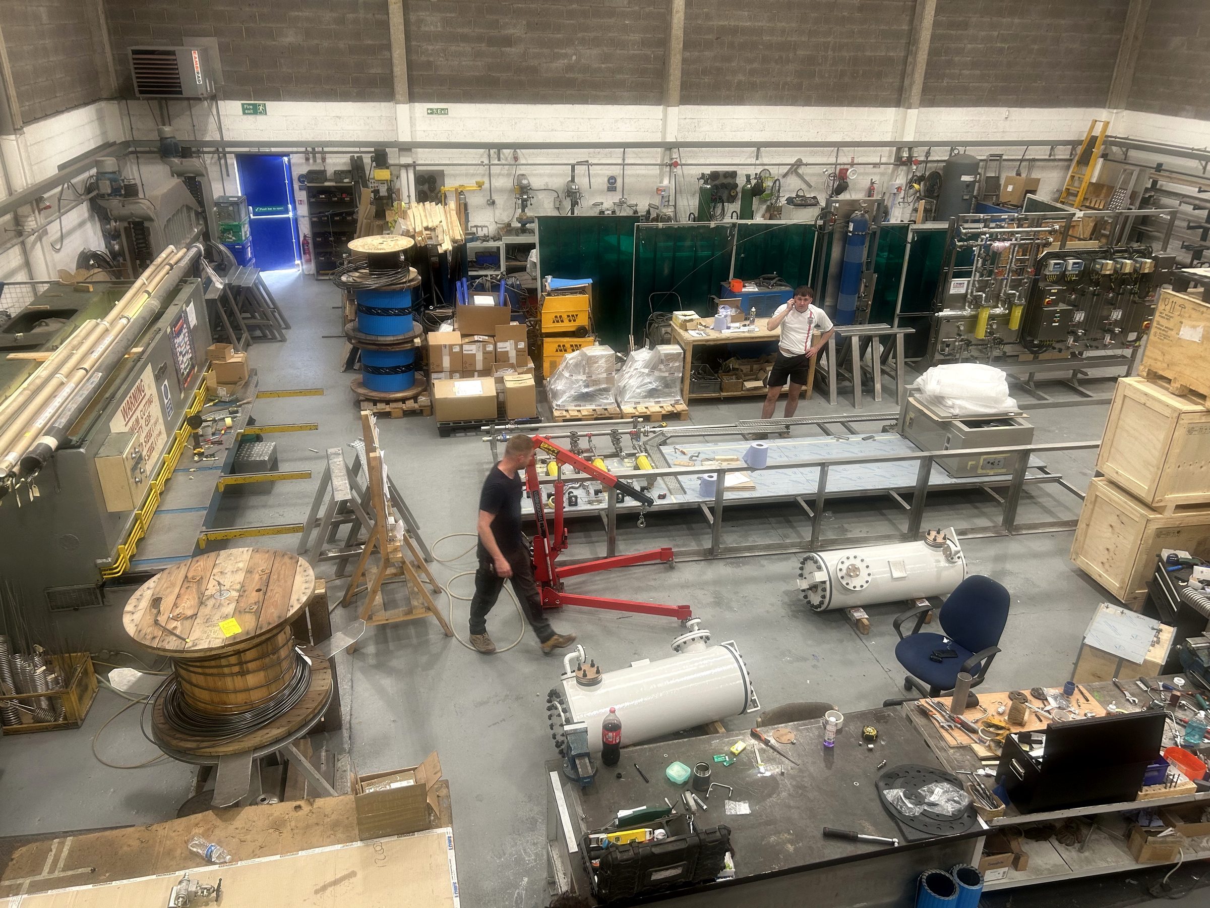 The workshop of an established UK engineering business during a working shift: fabricated pressure vessels, cable drums and crated assemblies on the floor, with two men at work.